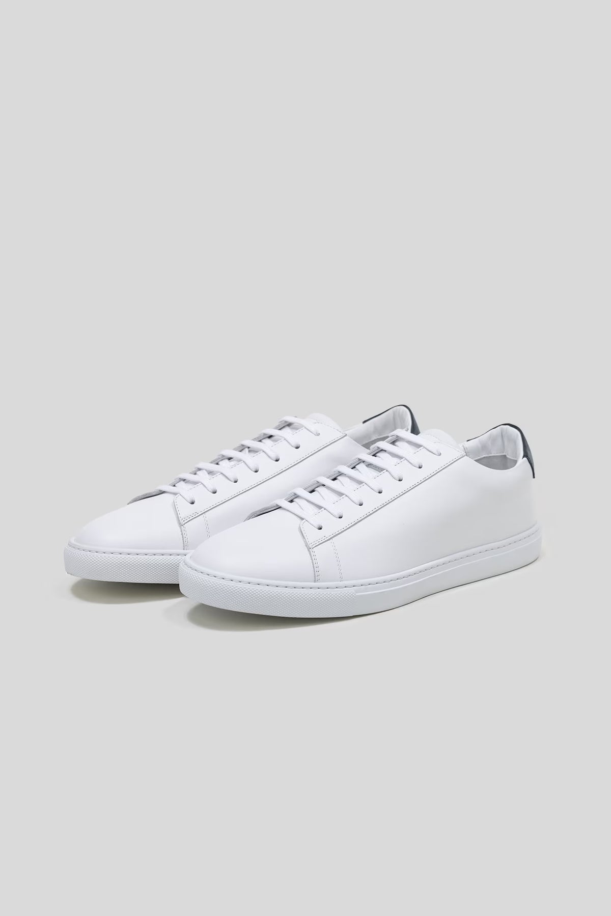 Three-quarter studio view of a pair of Olen Città low-top sneakers in white calfskin leather with navy blue heel tabs.