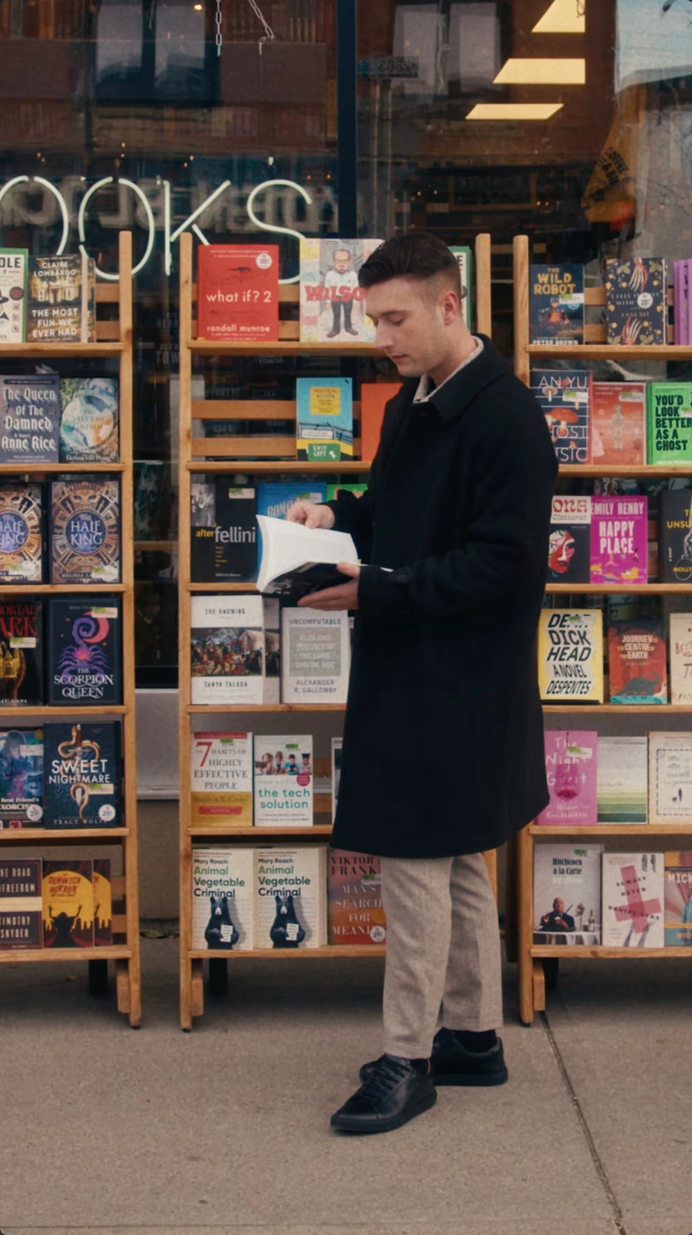 Man wearing a black coat and black calfskin Olen Città sneakers reading a book outside a bookstore window display.