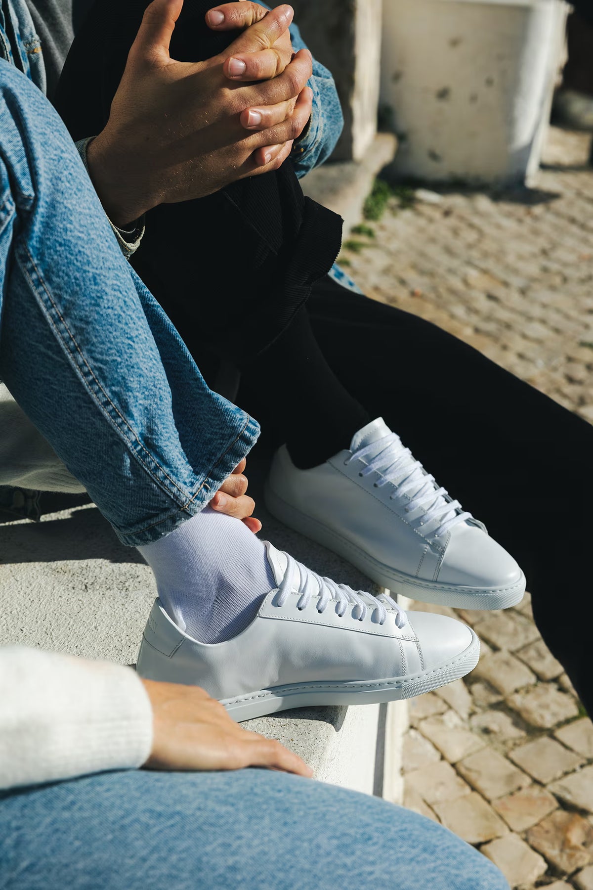 Close-up shot of two pairs of Olen Città low-top sneakers in white calfskin leather with white laces and cup soles, worn on a cobblestone street.