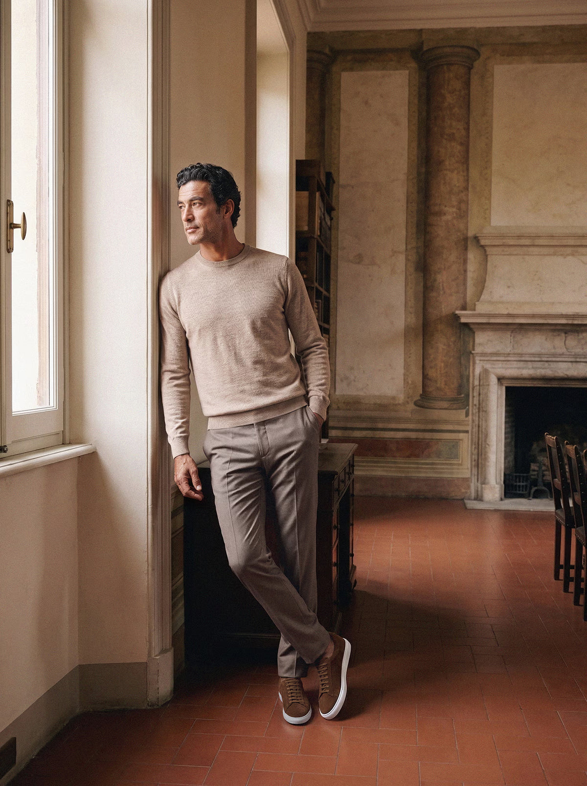 Man in a beige crewneck sweater, taupe trousers and tan suede sneakers leaning against a sunlit window frame
