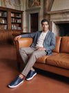 Man in a blue-gray blazer, cream knit polo, taupe trousers and navy leather sneakers seated on a tan leather sofa
