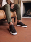 Dark brown leather low-top sneakers with white soles, worn with olive chinos while seated on a tan leather chair