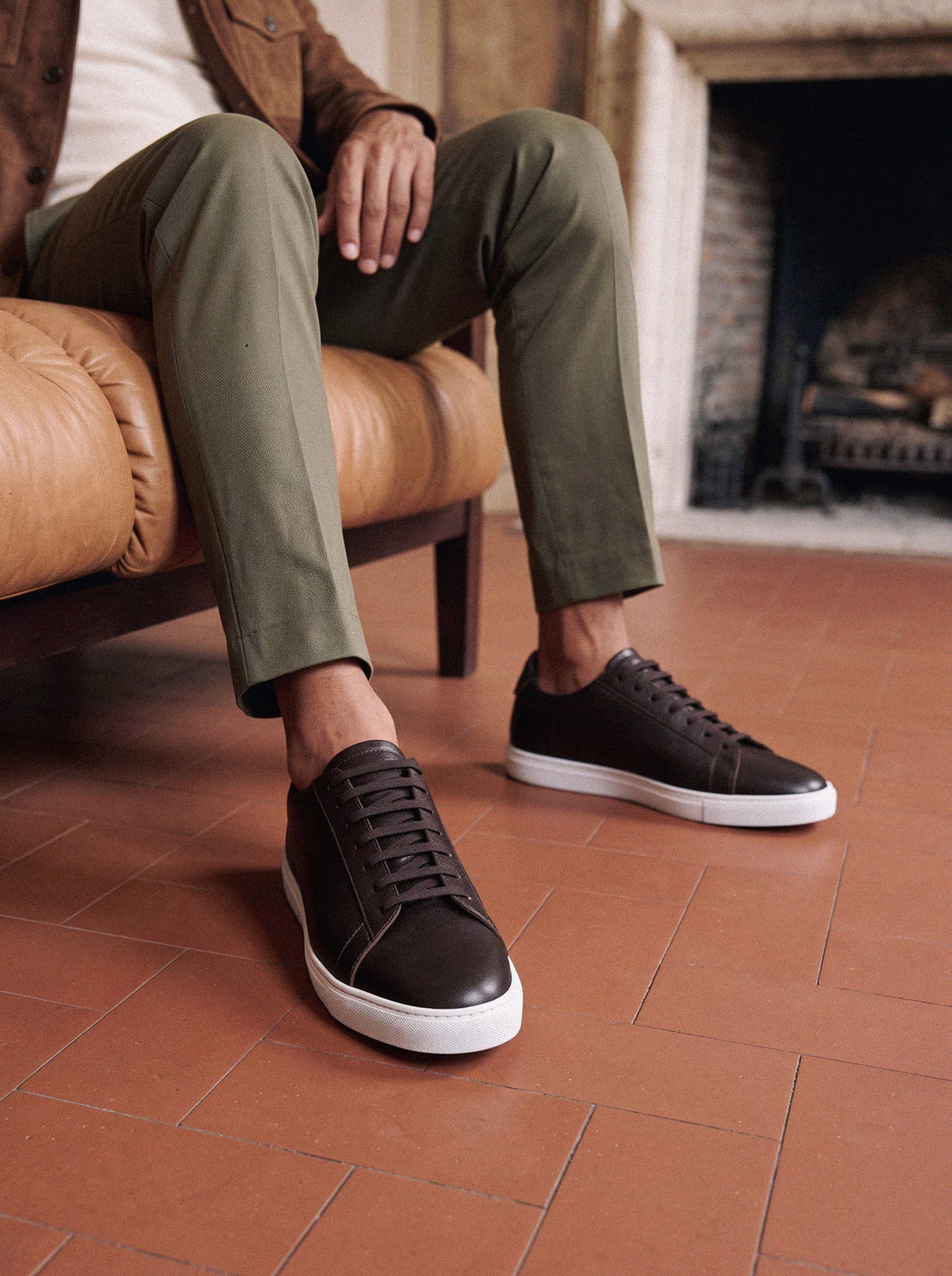 Dark brown leather low-top sneakers with white soles, worn with olive chinos while seated on a tan leather chair