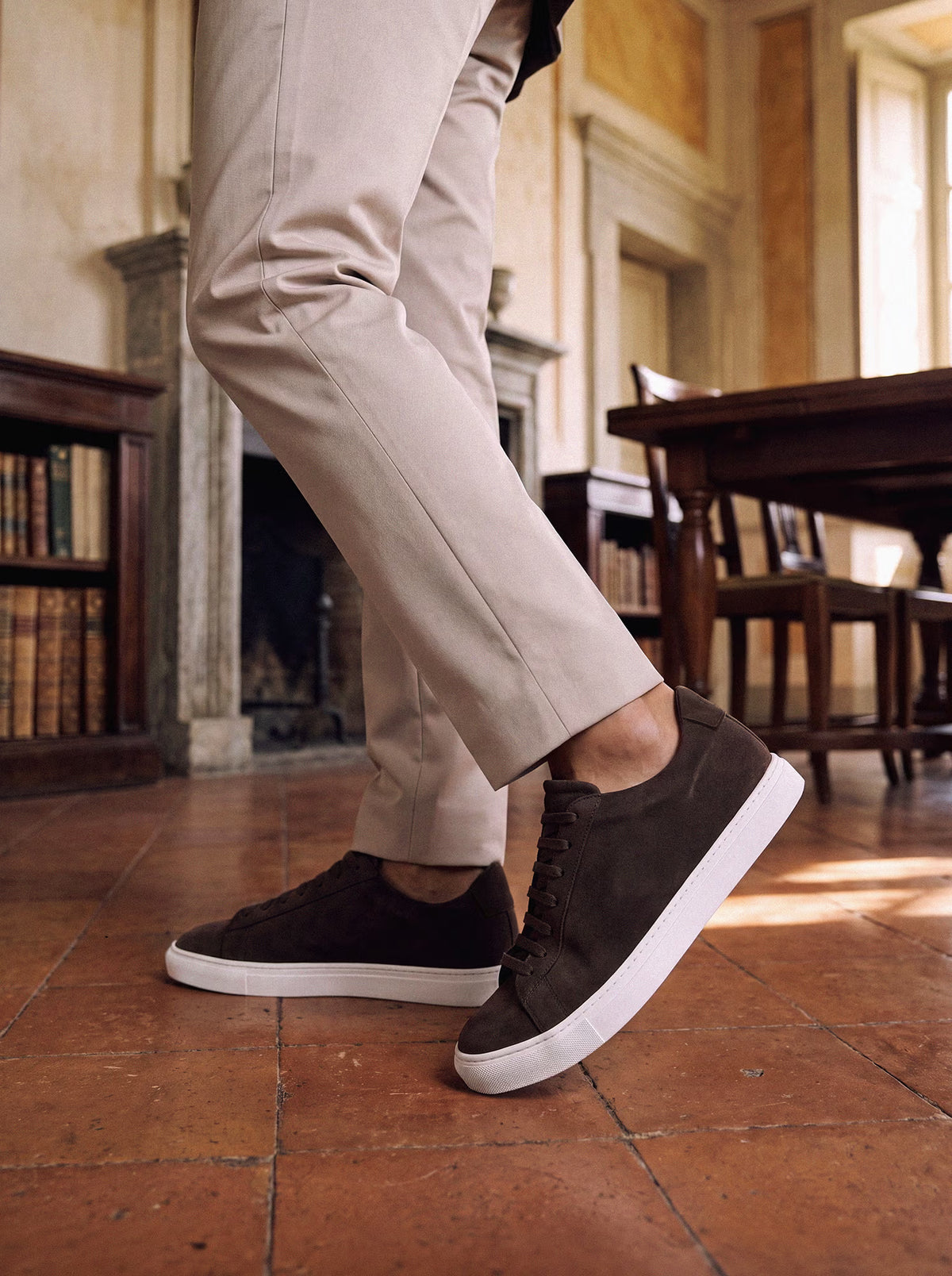 Dark brown suede low-top sneakers with white soles, worn with beige chinos in a classic library interior