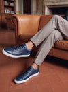 Navy blue leather low-top sneakers with white soles, worn with taupe cuffed trousers while seated on a leather sofa