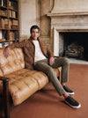 Man wearing a tan suede overshirt, cream crewneck sweater, olive chinos and brown leather sneakers on a tan leather sofa