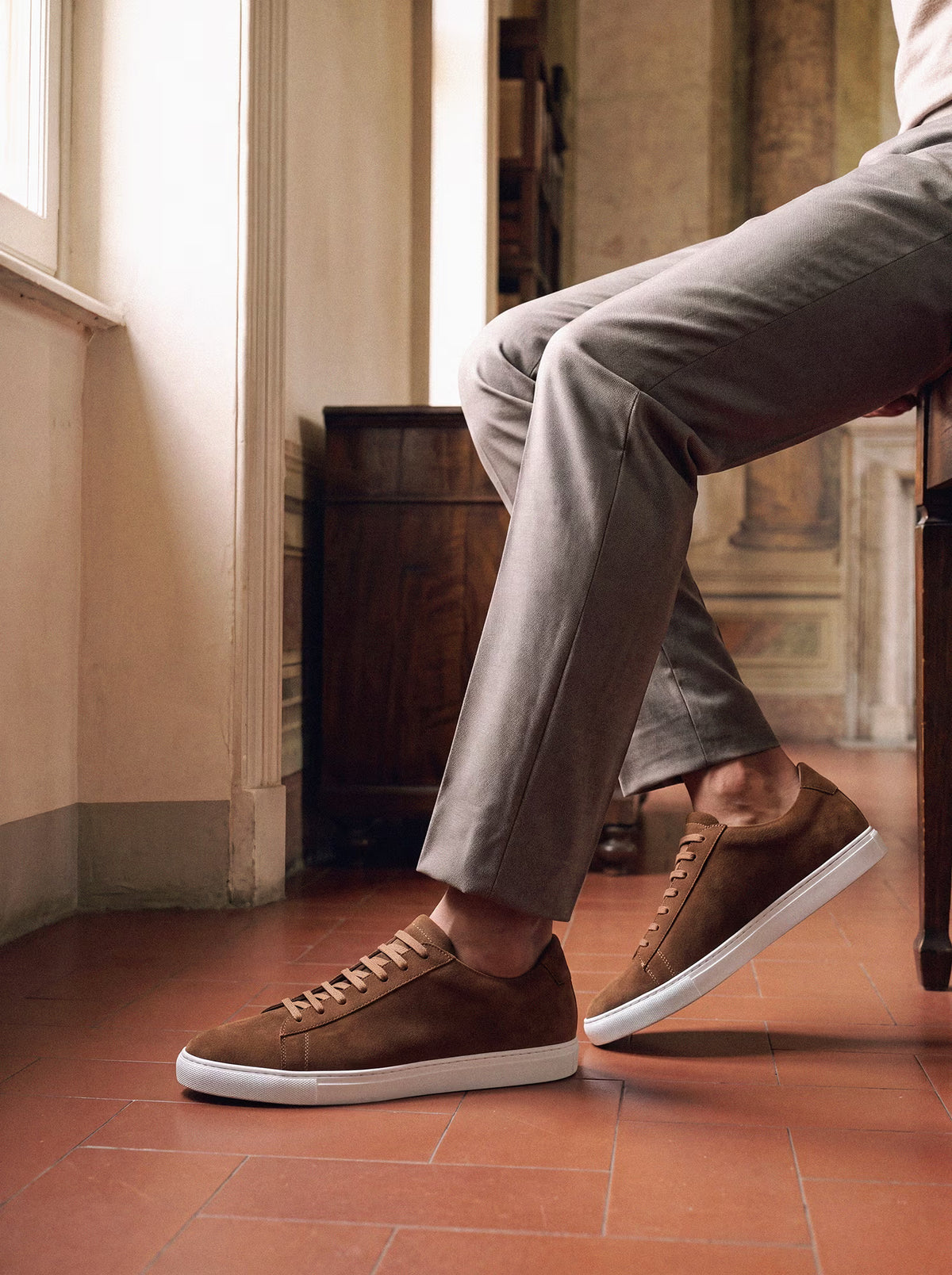 Tan suede low-top sneakers with white soles, worn with light gray trousers in a sunlit classic interior