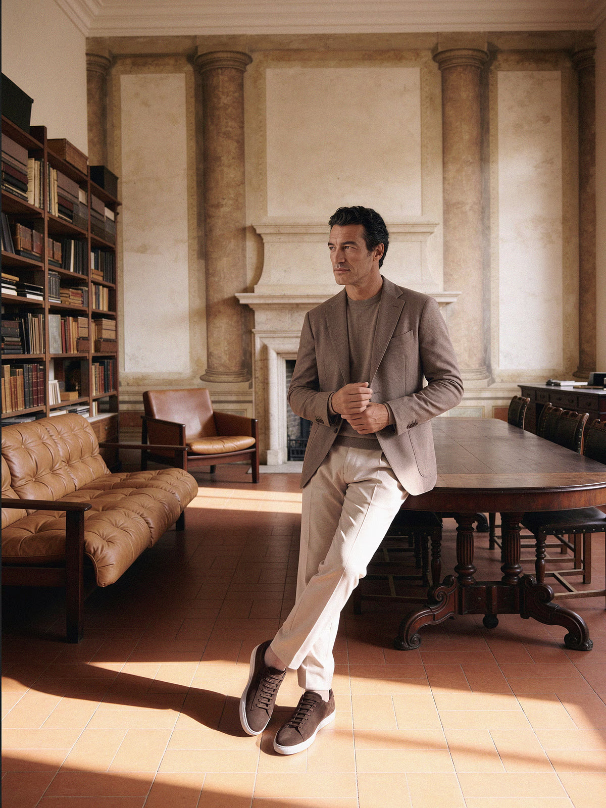Man in a taupe unstructured blazer, beige chinos and dark brown suede sneakers leaning on a wooden table in a library