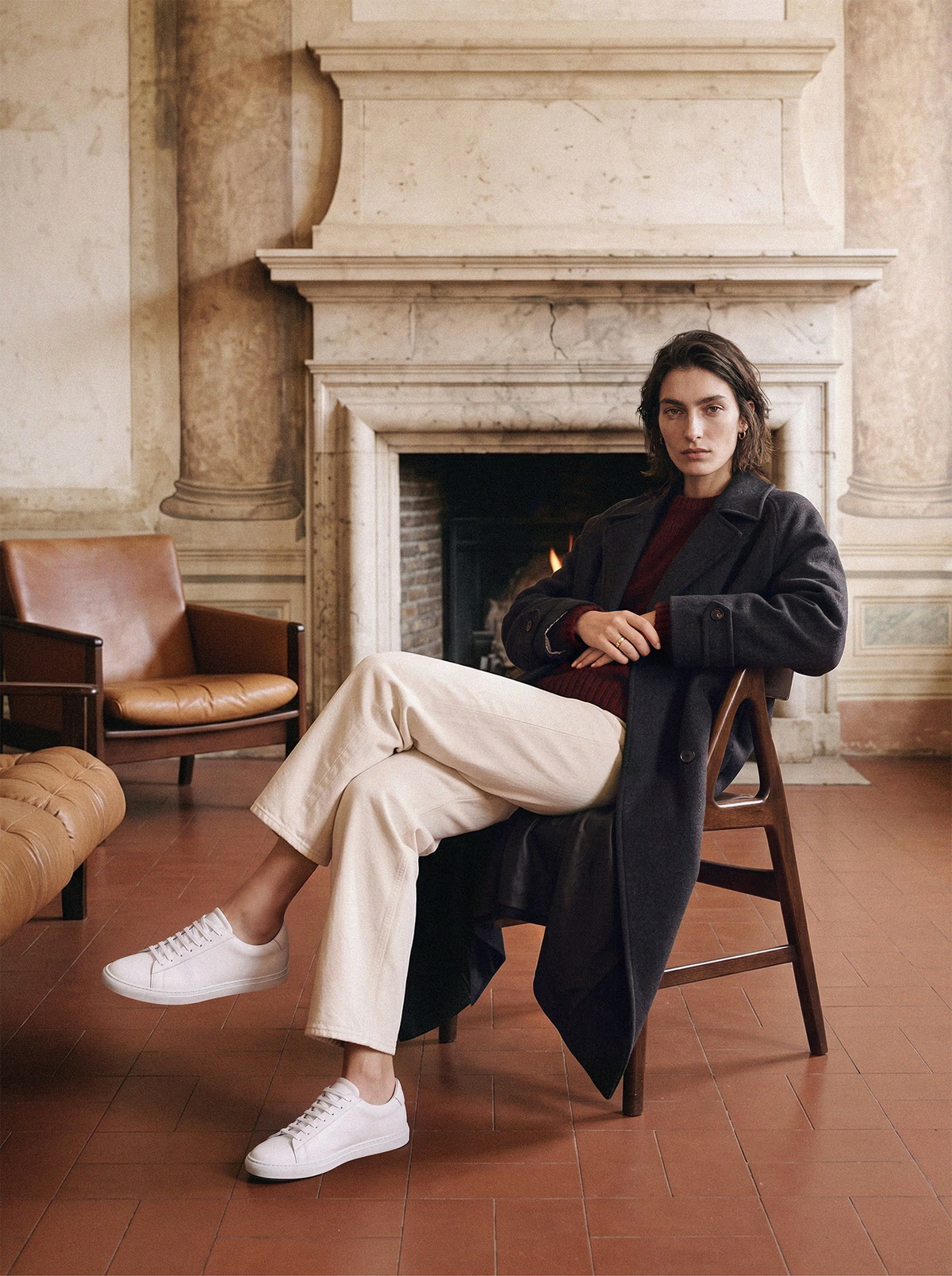 Woman in a charcoal wool coat, cream jeans and white leather sneakers seated on a wooden chair by a stone fireplace